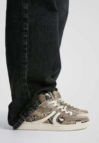 Baskets montantes avec un motif texturé de peau de serpent beige et marron, lacets blancs, semelle en caoutchouc crème, associées à un jean noir en denim.