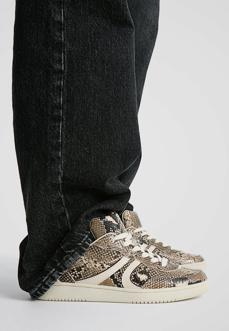 Baskets montantes avec un motif texturé de peau de serpent beige et marron, lacets blancs, semelle en caoutchouc crème, associées à un jean noir en denim.