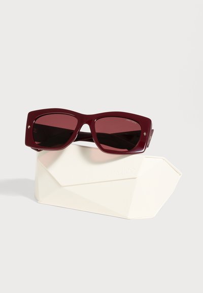 Des lunettes de soleil Jimmy Choo bordeaux aux verres foncés reposent sur un étui blanc siglé, sur un fond clair et uni.
