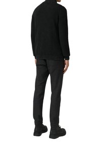 Mann in strukturiertem schwarzen Pullover, dunklen grauen Hosen und schwarzen Stiefeln, der von hinten gegen einen weißen Hintergrund steht.