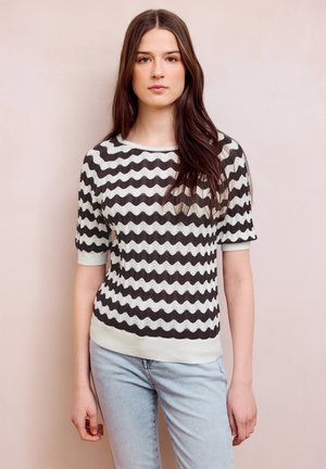 Mladá žena s dlouhými hnědými vlasy, oblečená v černo-bílém svetr s vlnitými pruhy a světlé modré džíny, stojící opřená o béžovou zeď.