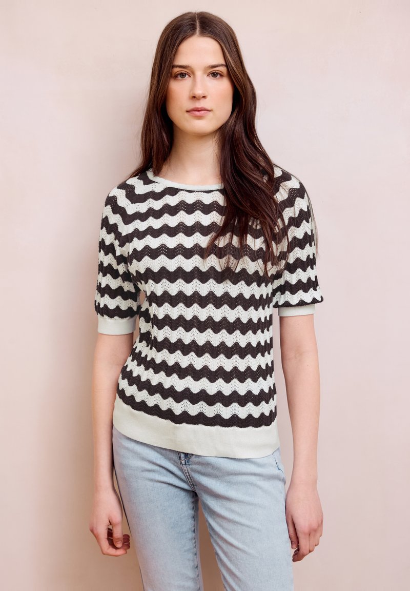 Jeune femme aux longs cheveux bruns portant un pull à manches courtes à rayures ondulées noir et blanc, et un jean bleu clair, debout contre un mur beige.