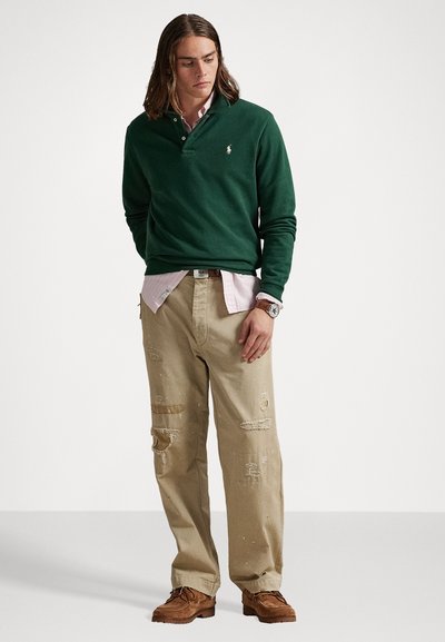Pull vert foncé avec un col, superposé à une chemise à col rose clair. Un jean beige déchiré et des chaussures marron complètent la tenue.