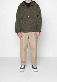 Personne portant un pull à capuche vert olive à motif craquelé, pantalon beige et baskets vertes et blanches, debout devant un fond blanc uni.