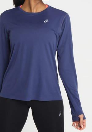 Frau trägt ein marineblaues Sportoberteil mit langen Ärmeln und schwarze Leggings mit ASICS-Logos, steht vor einem einfarbigen Hintergrund.