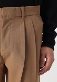 Bruna skräddarsydda byxor med flat front, veck och sidofickor. Tyget verkar slätt med en strukturerad passform vid midjan.