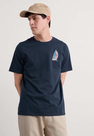Marineblauwe katoenen T-shirt met een kleine zeilbootafbeelding in rood, blauw en wit, met een ronde hals en korte mouwen. Beigekleurige pet gedragen.