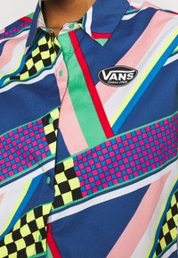Chemise Vans colorée avec des rayures diagonales en bleu, rose et vert. Présente des motifs à carreaux et un logo circulaire Vans sur la poitrine.