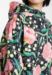 Kapuzensweatshirt mit schwarzem Hintergrund, versehen mit großen pinkfarbenen Blumen und grünen Blättern, akzentuiert durch kleine gelbe Blumenmuster.