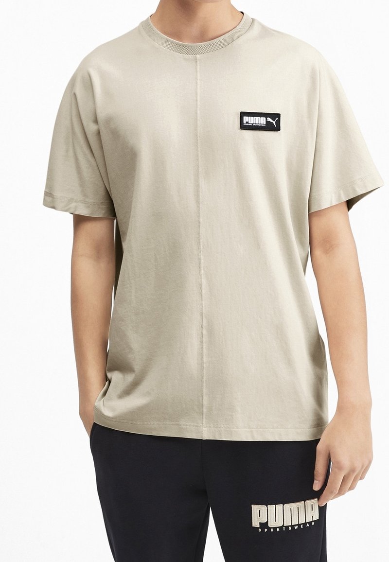 Beige bavlněné tričko s krátkým rukávem, kulatým výstřihem a černou obdélníkovou nášivkou loga PUMA na přední straně, spárované s černými joggingovými kalhotami.