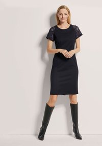 Schwarzes Kleid mit kurzen Spitzenärmeln und figurbetonter Silhouette, kombiniert mit knielangen schwarzen Lederstiefeln. Glatte Textur, knielanger Schnitt.