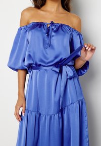 Robe en satin bleu décolletée avec un col à nouer, des manches bouffantes et une taille ceinturée avec un ourlet à volants. Texture lisse.