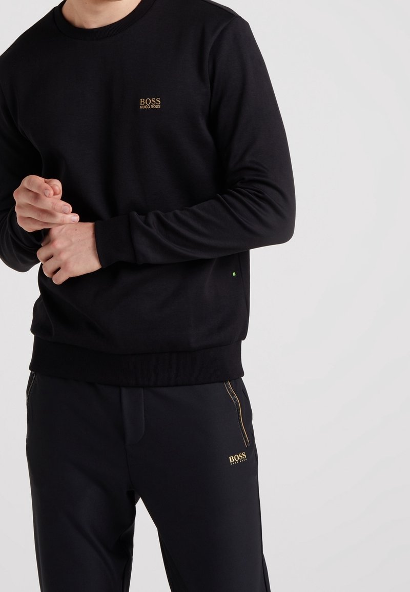 Svart sweatshirt med ribbade muddar och fåll, prydd med en liten gul logotyp på bröstet och en subtil grön accent längst ner. Matchas med svarta byxor.