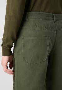 Olive green cotton pants with a textured finish. Features a back pocket and waistband detail, shown from the side with a close-up perspective.