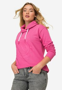 Felpa rosa con cappuccio, caratterizzata da un logo testurizzato sul fronte, con cordini e maniche lunghe. Indossata con jeans grigi, mani in tasca.