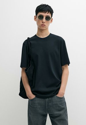 Person mit runder Sonnenbrille, schwarzem T-Shirt und grauen Jeans, mit einer Hand in der Tasche und einer schwarzen Tasche über der Schulter, steht vor einem schlichten Hintergrund.