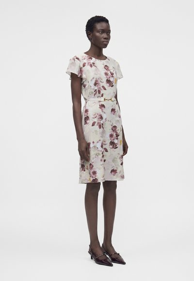 Γυναίκα που φοράει ένα λευκό φόρεμα μέχρι το γόνατο με ροζ και μοβ λουλουδάτα σχέδια, λεπτή ζώνη και μπορντό μυτερά τακούνια, στέκεται μπροστά σε απλό φόντο.