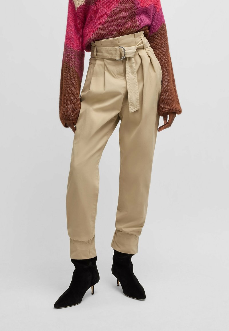 Pantalon beige taille haute ceinturé style paperbag, associé à un pull en maille multicolore et des bottines noires à bout pointu.