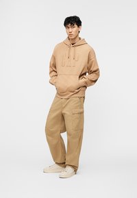 Νεαρός άνδρας που στέκεται με τα χέρια στις τσέπες, φοράει γκριζωπή μπουφάν GAP, φαρδιά cargo παντελόνια και κρεμ αθλητικά παπούτσια, μπροστά σε απλό φωτεινό φόντο.