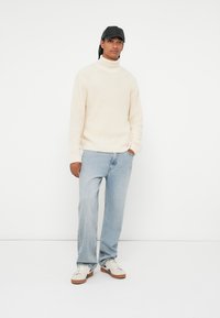 Maglione a collo alto a maglia, jeans dritti azzurri chiari, sneakers bianche con suole marroni e un berretto nero indossato dritto.