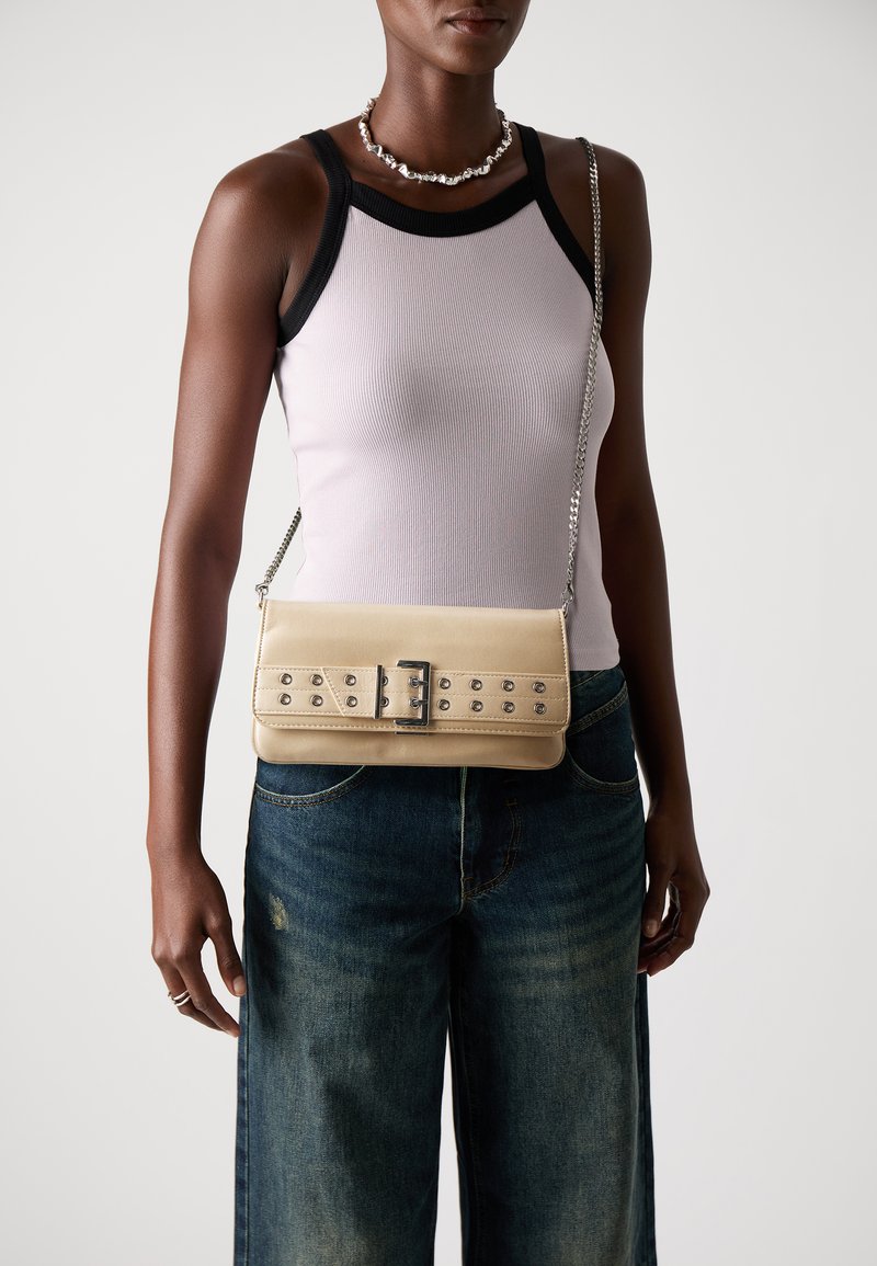 Sac à main beige avec une bandoulière en chaîne, arborant un design de ceinture cloutée et une boucle en argent. Texture lisse, forme rectangulaire. Porté en bandoulière.
