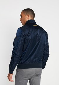 Navy blue bomber jacket with a shiny satin finish, ribbed cuffs, and a zippered pocket on the left sleeve. Features yellow text on the back.