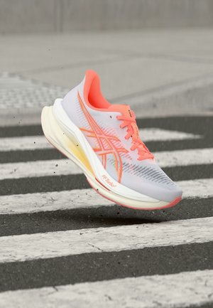 Chaussure de course gris clair et rose avec des lacets orange en l'air au-dessus d'un passage piéton sur une route asphaltée.