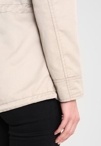 Beżowy rękaw kurtki z widocznym detalem szwów, wykonany z trwałego materiału. Ręka spoczywa na czarnych spodniach, podkreślając kontrastujące kolory.