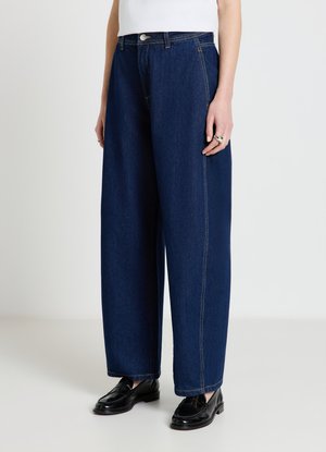 Personne portant un jean bleu foncé à jambes larges, des mocassins noirs et un haut blanc, debout devant un fond clair uni.