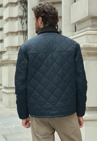 Padded Jacke in dunkelblau mit Rautensteppung, schwarzem Kragen und langen Ärmeln, getragen über hellbeigen Hosen.