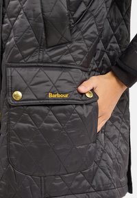 Zwarte gewatteerde vest met een grote voorkantzak. Gouden hardware-accenten en "Barbour" in gouden stiksel op de zak. Gladde textuur.