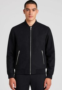 Uomo che indossa una giacca bomber nera con zip e due tasche con zip, in piedi contro uno sfondo grigio chiaro.