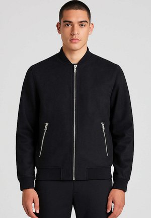 Uomo che indossa una giacca bomber nera con zip e due tasche con zip, in piedi contro uno sfondo grigio chiaro.