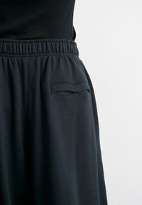 Midjan och övre låret på en person som bär svarta byxor med elastisk midja och en enkel slitsficka på höger sida, mot en enfärgad vit bakgrund.