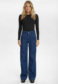 Tamsiai mėlynos aukštu liemeniu džinsai su laisvu kirpimu, turintys mygtukų užtrauktuką ir penkias kišenes, derinami su prigludusia juoda ilgomis rankovėmis palaidine.