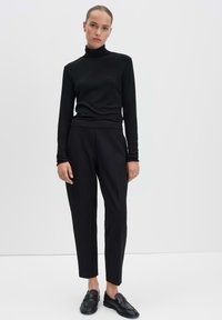 Zwart coltrui gecombineerd met nauwsluitende zwarte broek, beide gemaakt van glad materiaal. Model draagt zwarte instappers en staat tegen een witte achtergrond.