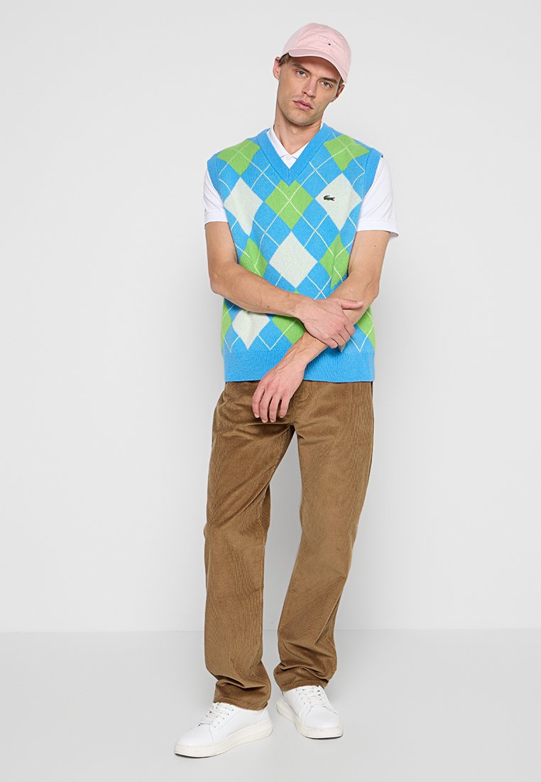 Un gilet en tricot à motif argyle bleu et vert sur une chemise blanche, associé à un pantalon en velours côtelé marron et des baskets blanches, le tout couronné d'une casquette rose.