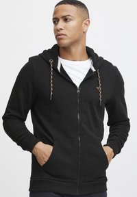 Sudadera con cremallera negra hecha de tejido suave, con cordón trenzado, bolsillos frontales y puños acanalados. Diseño sencillo con acentos mínimos.