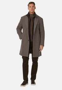 Cappotto di lana marrone con rever classico, chiusura a un bottone e due tasche frontali, indossato sopra un maglione scuro e pantaloni in corduroy marroni.