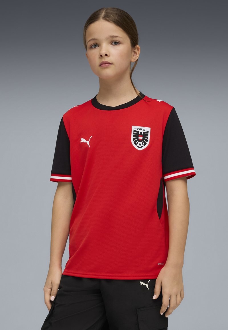 Κόκκινη και μαύρη ποδοσφαιρική φανέλα με κοντά μανίκια και στρογγυλή λαιμόκοψη, με λογότυπο στο στήθος και υφασμάτινη υφή. Διακοσμημένη με λευκές ρίγες.