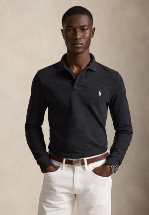 Hombre con polo de manga larga oscuro con logo bordado en blanco, pantalones blancos, cinturón marrón y reloj, de pie con las manos en los bolsillos.