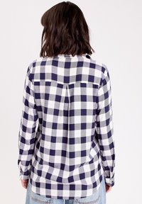 Chemise à manches longues à carreaux bleu marine et blanc avec un empiècement au dos, présentant un pli et des poignets à boutons. Le tissu a une texture douce.