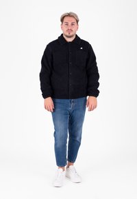 Zwarte fleece jack met knoopsluiting, twee voorzakken en gestructureerde stof. Draagt met blauwe spijkerbroek en witte sneakers.