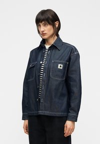 Σκούρο μπουφάν τζην με γιακά, δύο μπροστινές τσέπες, λευκές ραφές και ένα λογότυπο στο αριστερό τσεπάκι. Φορεμένο πάνω από πουκάμισο με ρίγες.