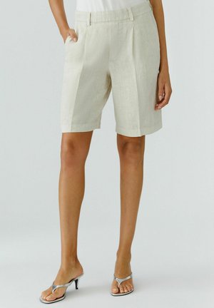 Frau trägt beige, knielange, maßgeschneiderte Shorts mit Taschen, kombiniert mit silbernen Sandalen mit Absatz, stehend vor einem schlichten Hintergrund.