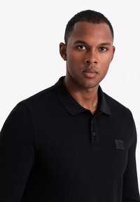 Svart stickad polo-shirt med långa ärmar, texturerad tyg och en liten rektangulär logotyp på bröstet. Klassisk krage och knappslå.