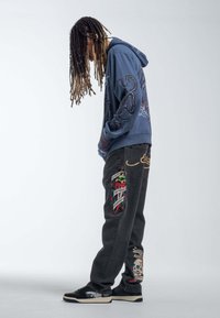 Hooded sweatshirt in blue with octopus graphic, paired with black jeans featuring colorful embroidered details, and black sneakers.