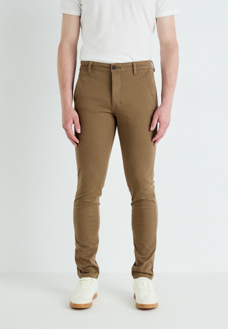 Hnědé slim-fit chinos vyrobené z pružné bavlny. Mají zapínání na knoflík, poutka na opasek a boční kapsy. Nosené s bílými teniskami.
