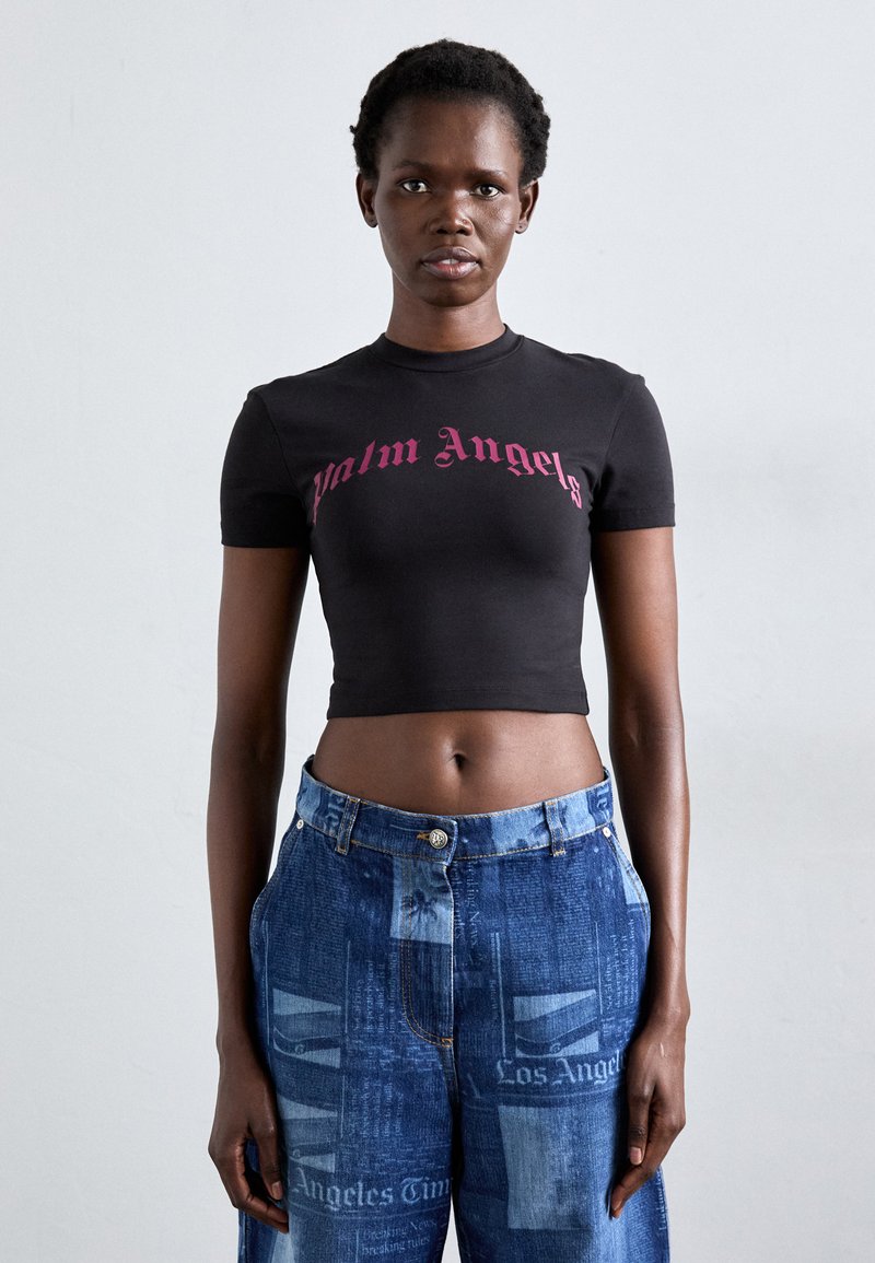 Woman wearing a black cropped t-shirt with "Palm Angels" text and blue jeans with newspaper print, standing against a plain light background.