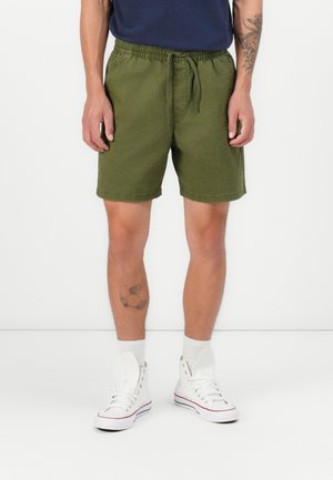 Shorts chinos verts avec une taille élastique et un cordon de serrage. Portés avec des baskets montantes blanches et des chaussettes visibles au niveau des chevilles.